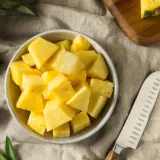 Frisch geschnittene Ananasstücke in einer Schüssel. Die süße Frucht, die manchmal leicht nach Alkohol schmeckt, liegt neben einem Messer und einem Stück Ananas auf einem Leinentuch.