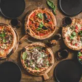 Vier verschiedene Pizzen, ein Salat und Rotwein auf Holztisch. Die Frage welcher Wein zu Pizza wird hier stilvoll beantwortet.