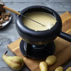 Ein dampfendes Schweizer Fondue in einem schwarzen Caquelon, daneben Brot, Kartoffeln und eine Birne.