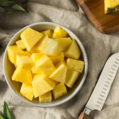 Frisch geschnittene Ananasstücke in einer Schüssel. Die süße Frucht, die manchmal leicht nach Alkohol schmeckt, liegt neben einem Messer und einem Stück Ananas auf einem Leinentuch.