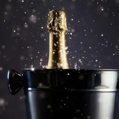 Goldener Champagnerflaschenhals mit Spritzern ragt aus einem Eiskübel. Das Champagner Etikett ist nicht sichtbar, aber die goldene Folie glänzt.