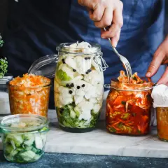 Fermentiertes Gemüse in Gläsern: Blumenkohl mit Pfefferkörnern, Karotten-Sauerkraut und ein Glas mit rotem Kimchi, das mit einer Gabel bearbeitet wird.