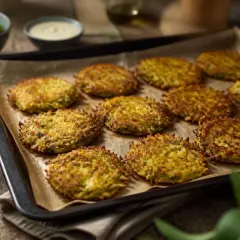 Goldbraune Kartoffel Zucchini Puffer auf einem Blech, bereit zum Genießen. Dazu ein Schälchen mit Dip und frischem Grün.