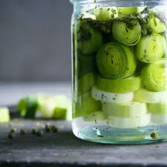 Ein Glas mit geschnittenem Lauch, bereit zum lauch fermentieren. Grüne Pfefferkörner und Kräuter sind ebenfalls zu sehen.