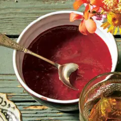 Ein Schälchen mit roter Soße, perfekt für ein Tkemali Rezept. Ein Löffel ruht darin, daneben ein Glas mit Bernstein-farbener Flüssigkeit und Blumen.