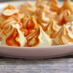 Orange-weiße Baiser nach dem Backen, die leicht klebrig sind. Sie liegen auf einer rosa Platte, die auf einem Holztisch steht.