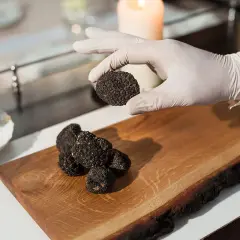 Ein Koch hält eine trufa negra über ein Holzbrett mit weiteren Exemplaren. Daneben steht eine Flasche Öl.