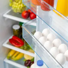 Ein Kühlschrank voller frischer Lebensmittel. In der Eierablage liegen viele weiße Eier, die noch gut sind. Daneben sind rote Paprika, Maiskolben und Trauben zu sehen.