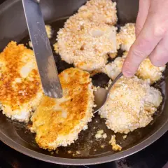 Goldbraune Blumenkohl-Schnitzel brutzeln in der Pfanne. Eine Hand wendet sie mit Messer und Löffel.