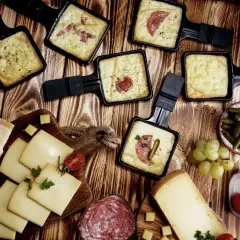 Raclette-Pfännchen mit geschmolzenem Käse, Brot, Käse, Salami, Cornichons und Trauben. Perfekt für einen gemütlichen Abend mit einem Glas Wein.