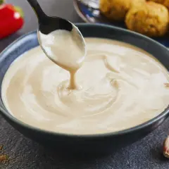 Cremige Tahini-Sauce tropft von einem Löffel in eine Schüssel. Sie ist die perfekte Begleitung zu Falafel, wie sie im Hintergrund zu sehen sind.