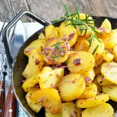 Goldbraune Bratkartoffeln mit Zwiebeln und Rosmarin in einer Pfanne, perfekt, um sie warm zu halten. Dazu liegen Gabel und Messer bereit.