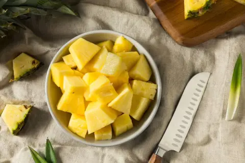 Frisch geschnittene Ananasstücke in einer Schüssel. Die süße Frucht, die manchmal leicht nach Alkohol schmeckt, liegt neben einem Messer und einem Stück Ananas auf einem Leinentuch.