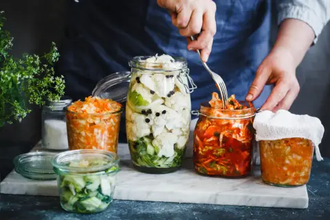 Fermentiertes Gemüse in Gläsern: Blumenkohl mit Pfefferkörnern, Karotten-Sauerkraut und ein Glas mit rotem Kimchi, das mit einer Gabel bearbeitet wird.