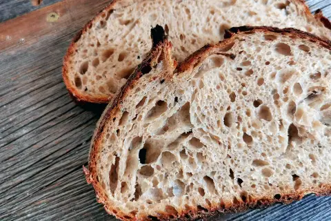 Zwei Scheiben rustikales, fermentiertes Brot liegen auf einer Holzoberfläche. Die Krume ist luftig und voller großer Poren.