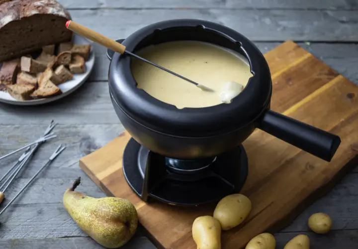 Ein dampfendes Schweizer Fondue in einem schwarzen Caquelon, daneben Brot, Kartoffeln und eine Birne.