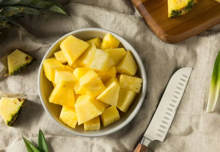 Frisch geschnittene Ananasstücke in einer Schüssel. Die süße Frucht, die manchmal leicht nach Alkohol schmeckt, liegt neben einem Messer und einem Stück Ananas auf einem Leinentuch.