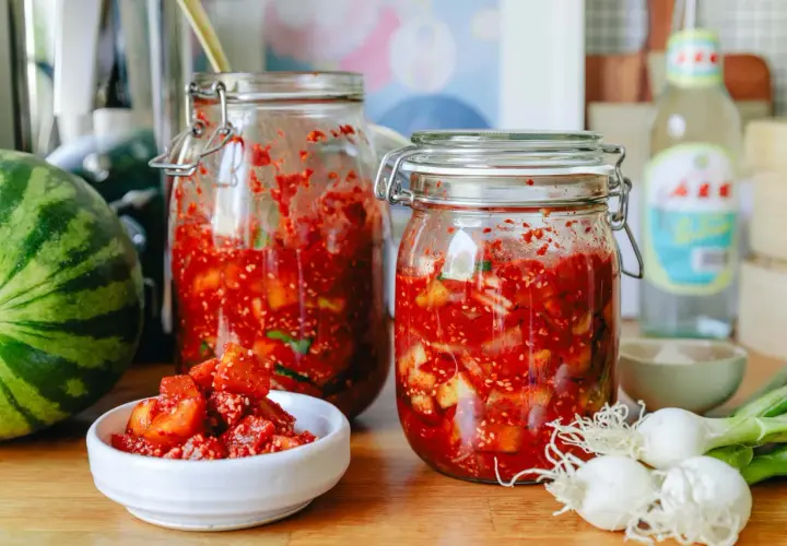 Zwei Gläser mit fermentierter Melone, daneben eine Wassermelone, Frühlingszwiebeln und eine Flasche.