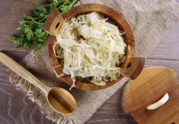 Holzschale mit Sauerkraut, daneben ein Holzlöffel und ein Stück Knoblauch. Manche finden, dass Sauerkraut schmeckt bitter.