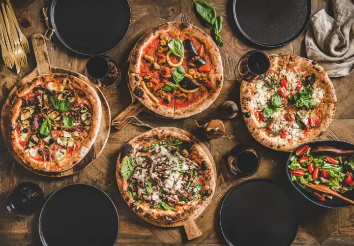 Vier verschiedene Pizzen, ein Salat und Rotwein auf Holztisch. Die Frage welcher Wein zu Pizza wird hier stilvoll beantwortet.