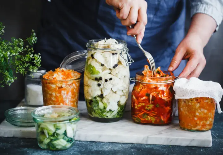 Fermentiertes Gemüse in Gläsern: Blumenkohl mit Pfefferkörnern, Karotten-Sauerkraut und ein Glas mit rotem Kimchi, das mit einer Gabel bearbeitet wird.