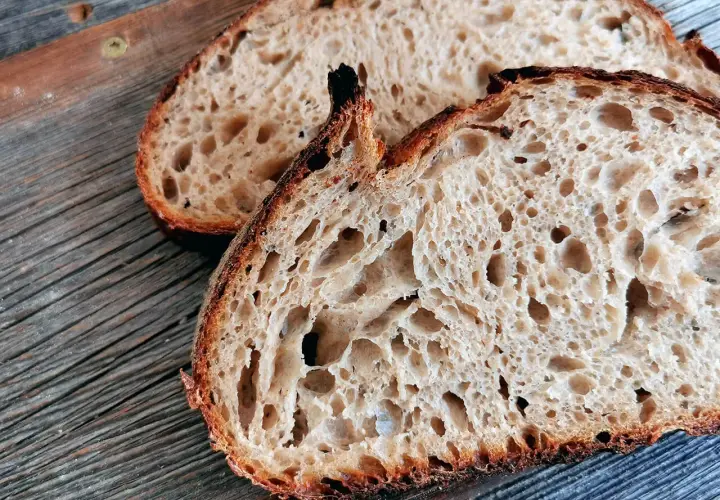 Zwei Scheiben rustikales, fermentiertes Brot liegen auf einer Holzoberfläche. Die Krume ist luftig und voller großer Poren.