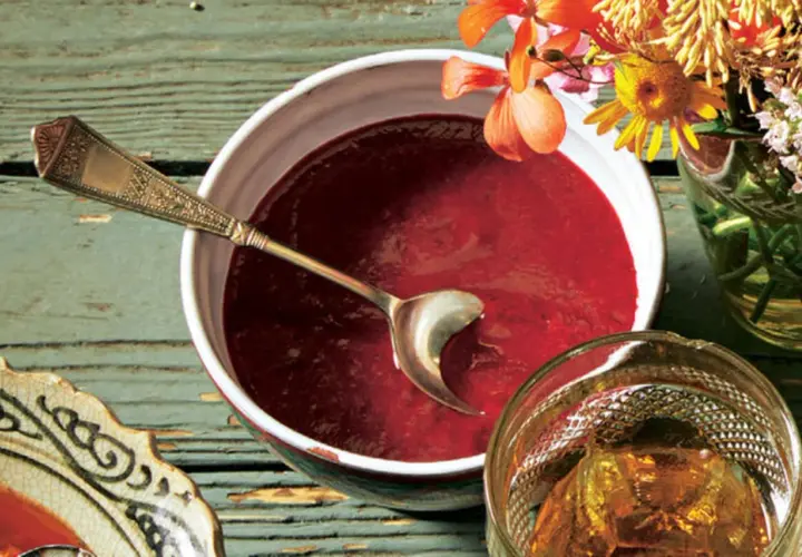 Ein Schälchen mit roter Soße, perfekt für ein Tkemali Rezept. Ein Löffel ruht darin, daneben ein Glas mit Bernstein-farbener Flüssigkeit und Blumen.