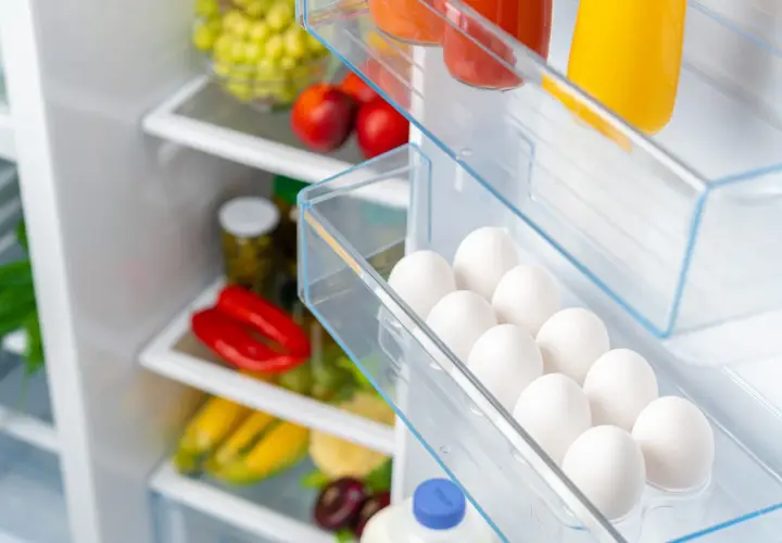 Ein Kühlschrank voller frischer Lebensmittel. In der Eierablage liegen viele weiße Eier, die noch gut sind. Daneben sind rote Paprika, Maiskolben und Trauben zu sehen.