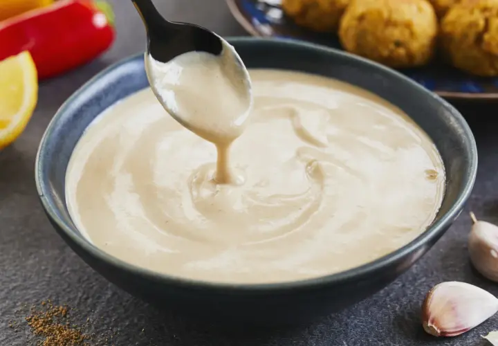 Cremige Tahini-Sauce tropft von einem Löffel in eine Schüssel. Sie ist die perfekte Begleitung zu Falafel, wie sie im Hintergrund zu sehen sind.