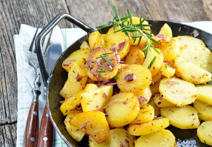 Goldbraune Bratkartoffeln mit Zwiebeln und Rosmarin in einer Pfanne, perfekt, um sie warm zu halten. Dazu liegen Gabel und Messer bereit.