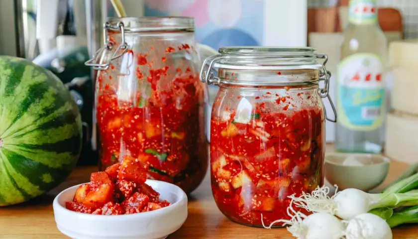 Zwei Gläser mit fermentierter Melone, daneben eine Wassermelone, Frühlingszwiebeln und eine Flasche.
