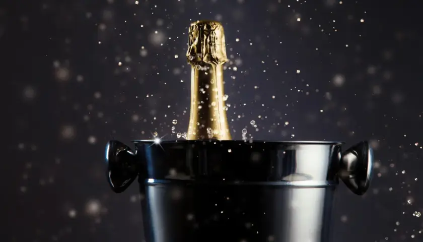 Goldener Champagnerflaschenhals mit Spritzern ragt aus einem Eiskübel. Das Champagner Etikett ist nicht sichtbar, aber die goldene Folie glänzt.