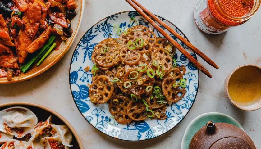 Knusprige, karamellisierte **Lotuswurzel**-Scheiben, garniert mit Frühlingszwiebeln, neben gedämpften Teigtaschen und einem Glas scharfer Chilisauce.