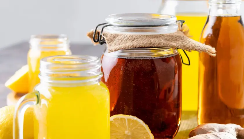 Gläser mit gelbem Getränk und ein großes Glas mit dunklem, sprudelndem fermentiertem Honig, daneben Zitronen und Ingwer.