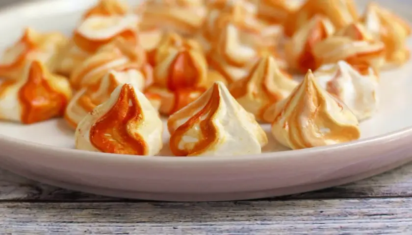 Orange-weiße Baiser nach dem Backen, die leicht klebrig sind. Sie liegen auf einer rosa Platte, die auf einem Holztisch steht.