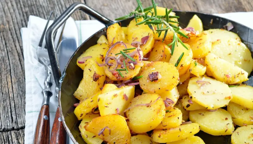 Goldbraune Bratkartoffeln mit Zwiebeln und Rosmarin in einer Pfanne, perfekt, um sie warm zu halten. Dazu liegen Gabel und Messer bereit.