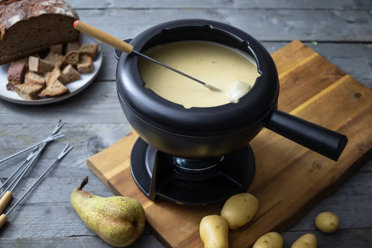 Ein dampfendes Schweizer Fondue in einem schwarzen Caquelon, daneben Brot, Kartoffeln und eine Birne.