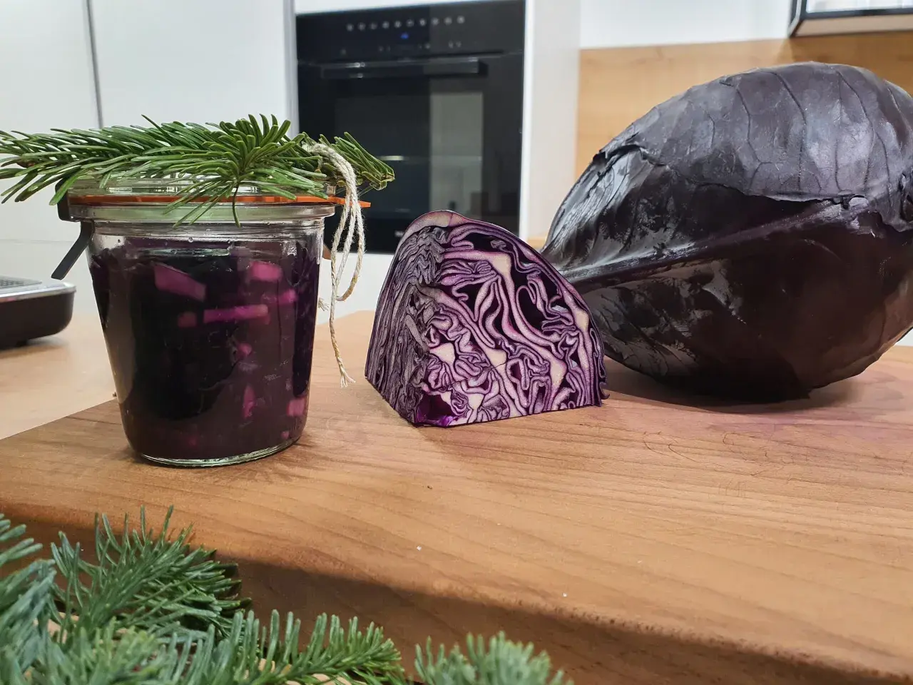 Ein Glas mit fermentiertem Rotkohl, daneben ein halber und ein ganzer Rotkohl. Tannenzweige schmücken das Glas.
