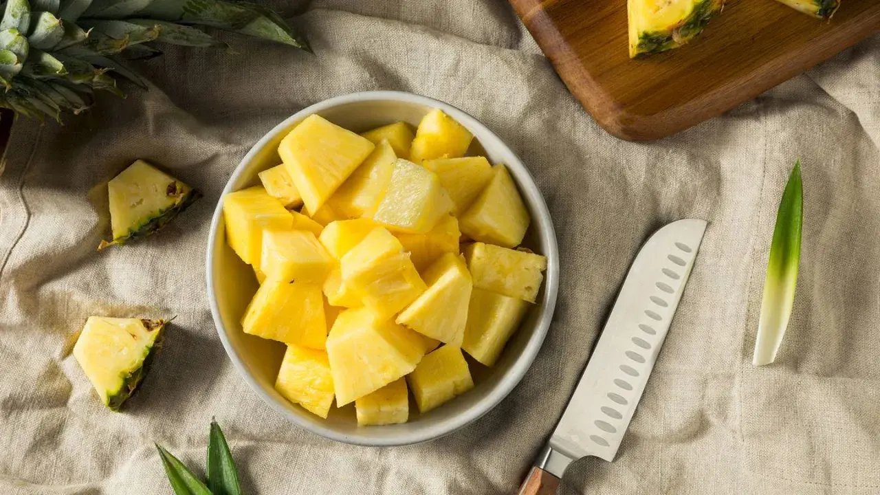 Frisch geschnittene Ananasstücke in einer Schüssel. Die süße Frucht, die manchmal leicht nach Alkohol schmeckt, liegt neben einem Messer und einem Stück Ananas auf einem Leinentuch.