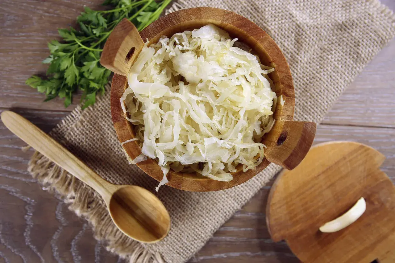 Holzschale mit Sauerkraut, daneben ein Holzlöffel und ein Stück Knoblauch. Manche finden, dass Sauerkraut schmeckt bitter.