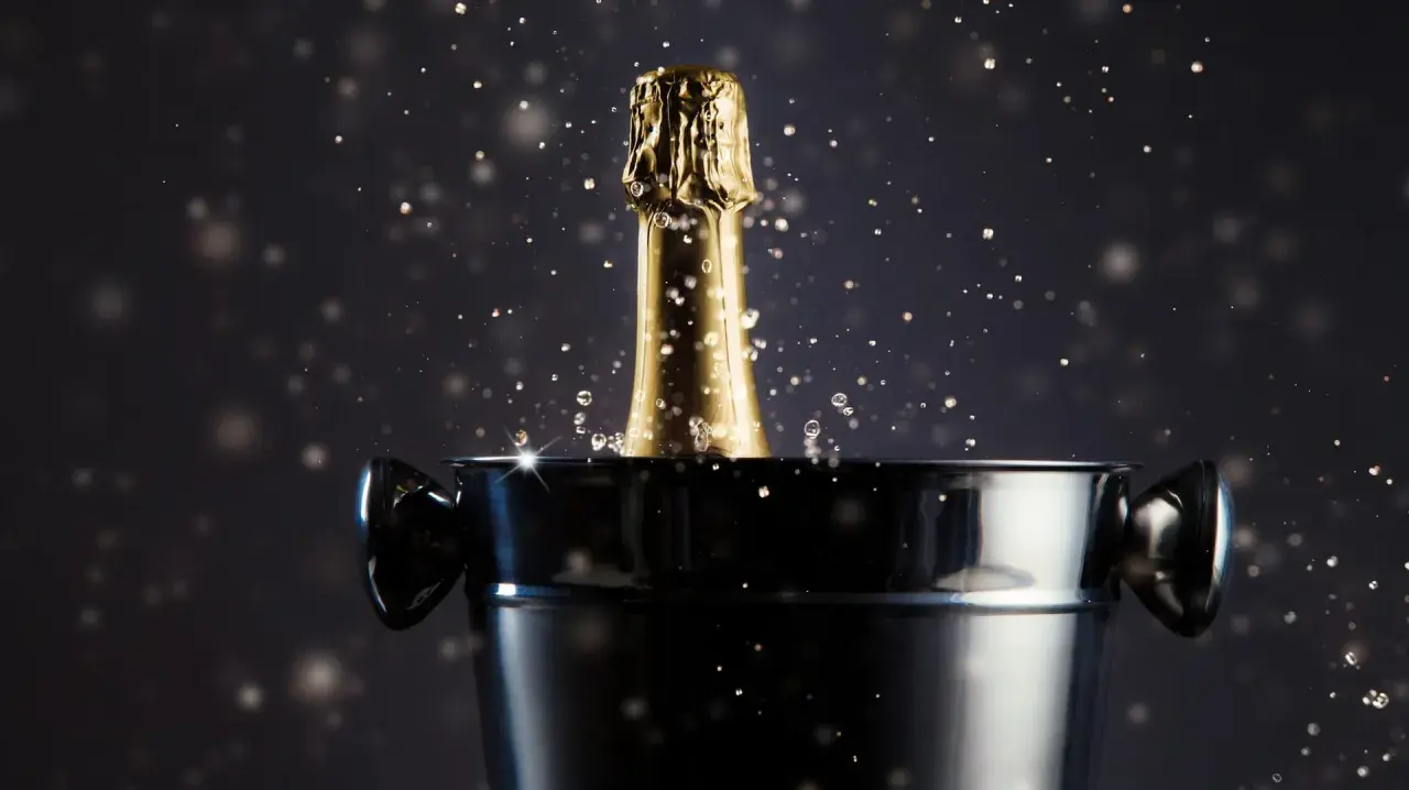 Goldener Champagnerflaschenhals mit Spritzern ragt aus einem Eiskübel. Das Champagner Etikett ist nicht sichtbar, aber die goldene Folie glänzt.