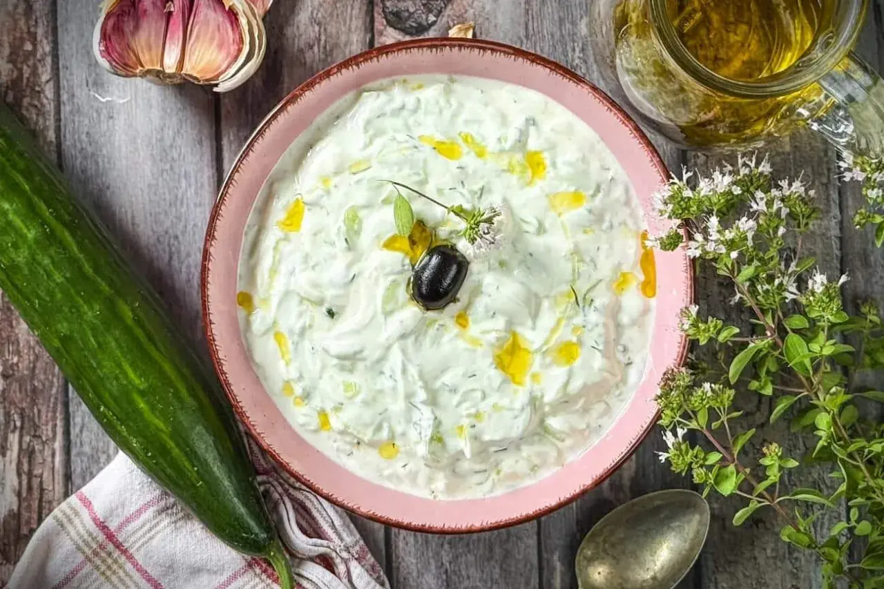 Schüssel mit cremigem griechischem Tzatziki, garniert mit Olive und Öl. Daneben Gurke, Knoblauch und frische Kräuter.