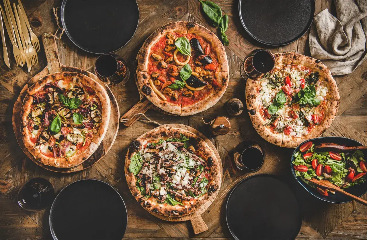 Vier verschiedene Pizzen, ein Salat und Rotwein auf Holztisch. Die Frage welcher Wein zu Pizza wird hier stilvoll beantwortet.