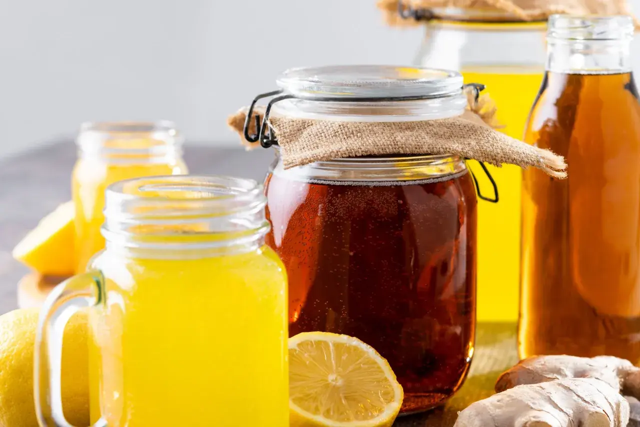 Gläser mit gelbem Getränk und ein großes Glas mit dunklem, sprudelndem fermentiertem Honig, daneben Zitronen und Ingwer.