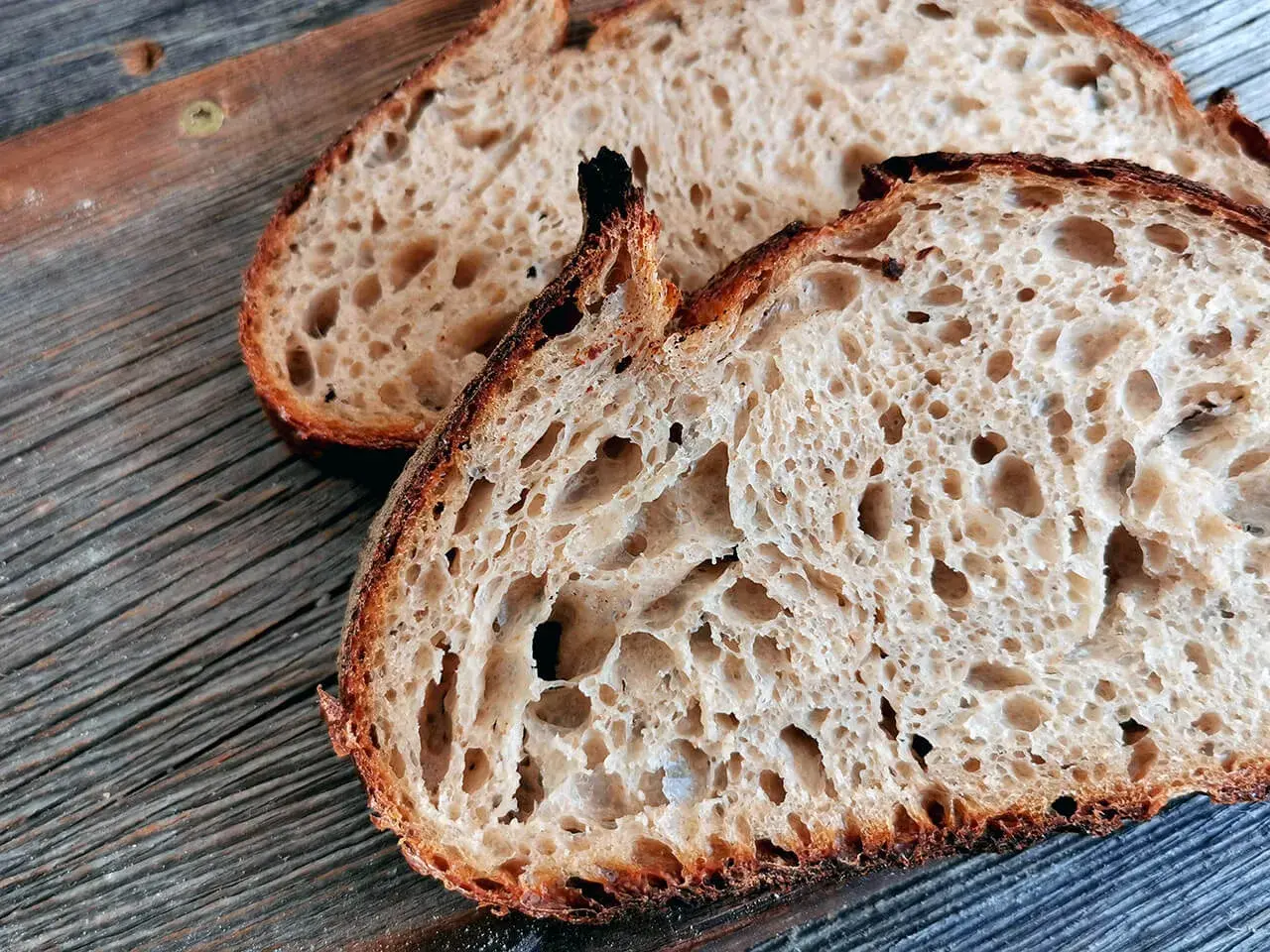 Zwei Scheiben rustikales, fermentiertes Brot liegen auf einer Holzoberfläche. Die Krume ist luftig und voller großer Poren.