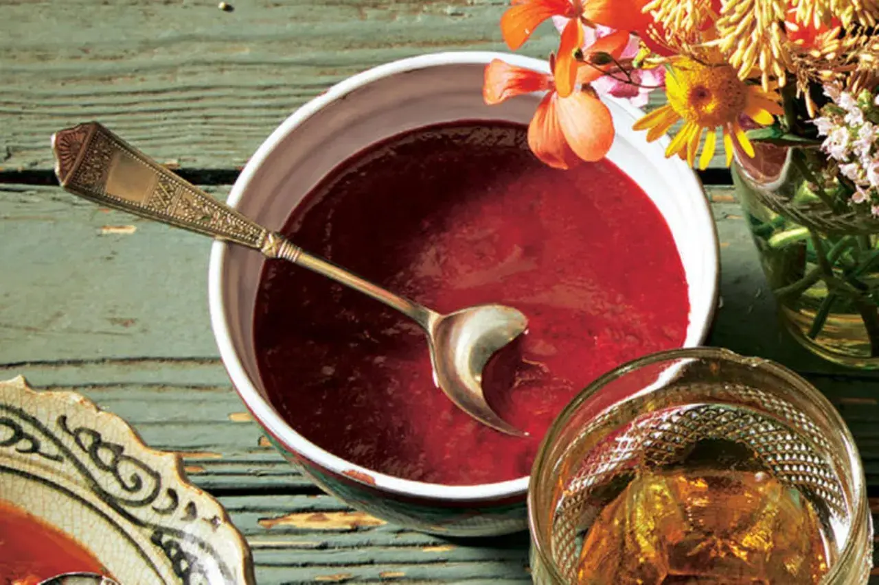 Ein Schälchen mit roter Soße, perfekt für ein Tkemali Rezept. Ein Löffel ruht darin, daneben ein Glas mit Bernstein-farbener Flüssigkeit und Blumen.