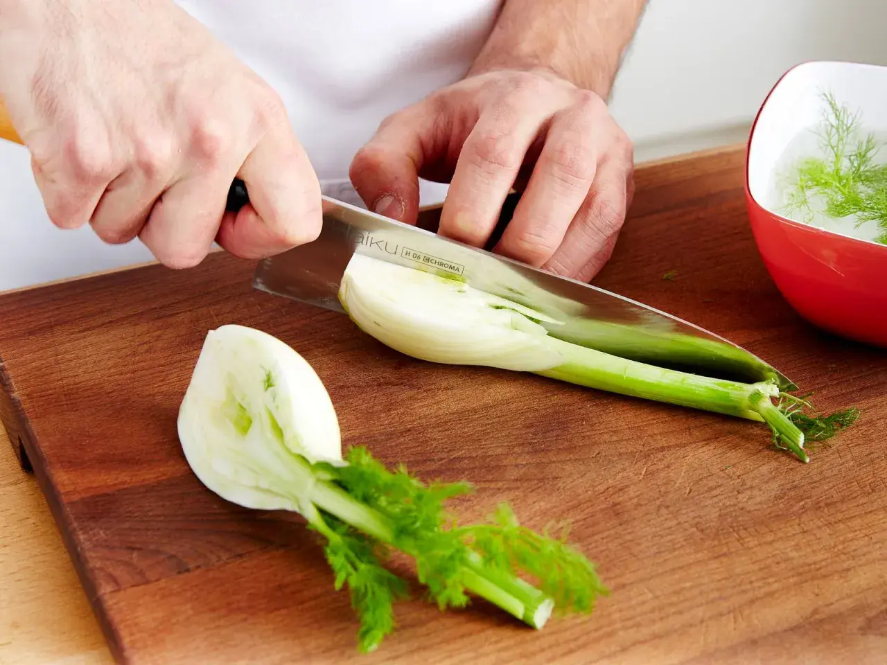 Hände schneiden Fenchel für ein Gericht mit feinem Fenchel Geschmack. Ein Teil liegt auf dem Holzbrett, ein anderer im roten Schälchen.