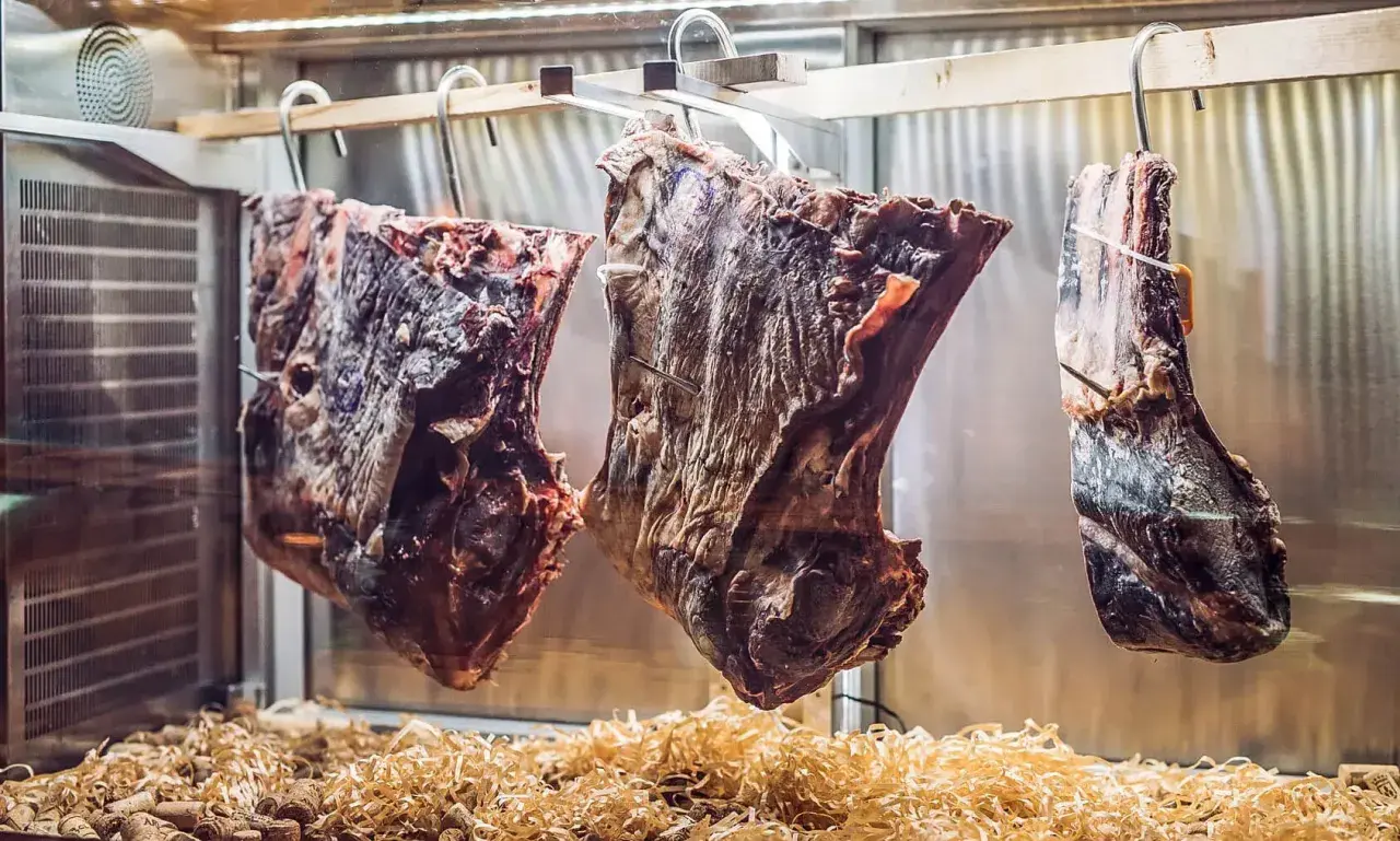 Drei große Fleischstücke hängen in einer Kühlkammer, bereit zum **Fleisch fermentieren**. Darunter liegen Holzspäne und Korken.