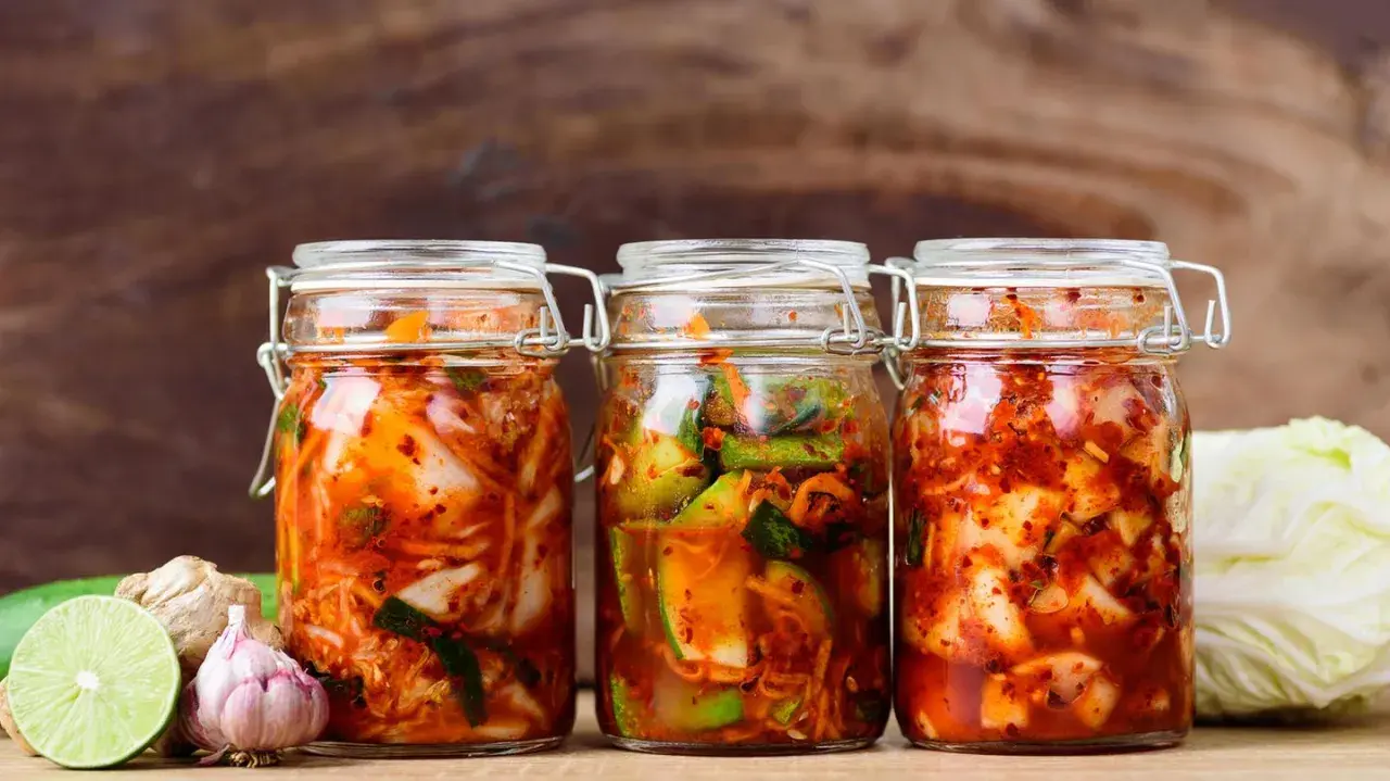 Drei Gläser mit koreanischer Vorspeise fermentierter scharfer Kohl (Kimchi) stehen bereit. Daneben liegen frische Zutaten wie Knoblauch, Ingwer und Limette.