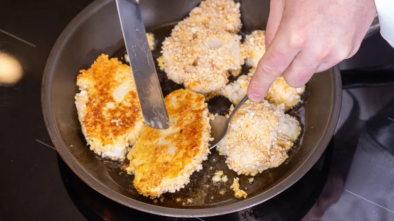 Goldbraune Blumenkohl-Schnitzel brutzeln in der Pfanne. Eine Hand wendet sie mit Messer und Löffel.